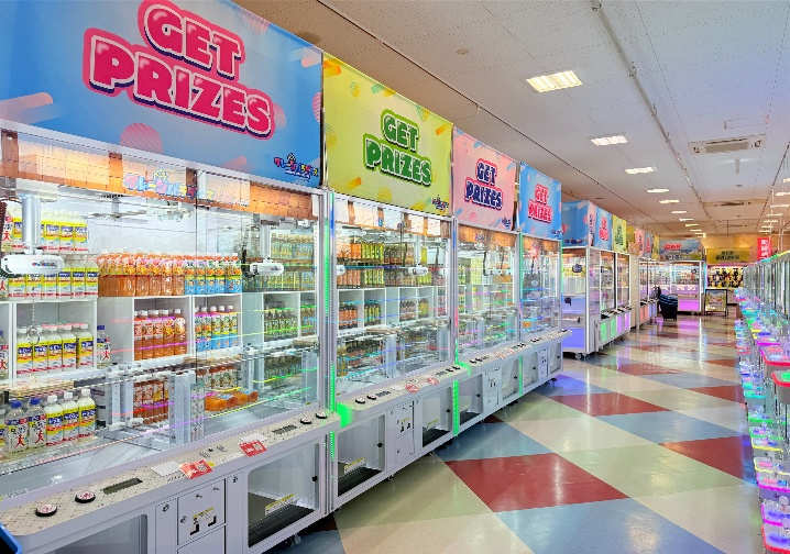 クレーンパラダイス諏訪店の店内。「GET PRIZES」のカラフルな看板が並ぶクレーンゲーム筐体の列で、景品はペットボトル飲料や日用品が中心。床は赤・青・白のチェッカー柄