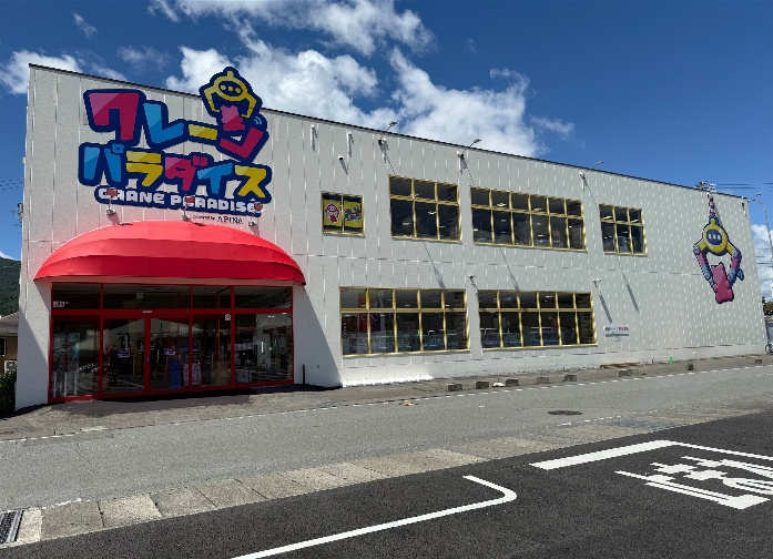 クレーンパラダイス諏訪店の外観。シルバーの外壁に「クレーンパラダイス CRANE PARADISE powered by APINA」の大型看板を掲げ、赤いアーチ型のひさしが付いたエントランスと黄色い窓枠、壁面にはアピナのマスコットキャラクター。背景は青空と山並み