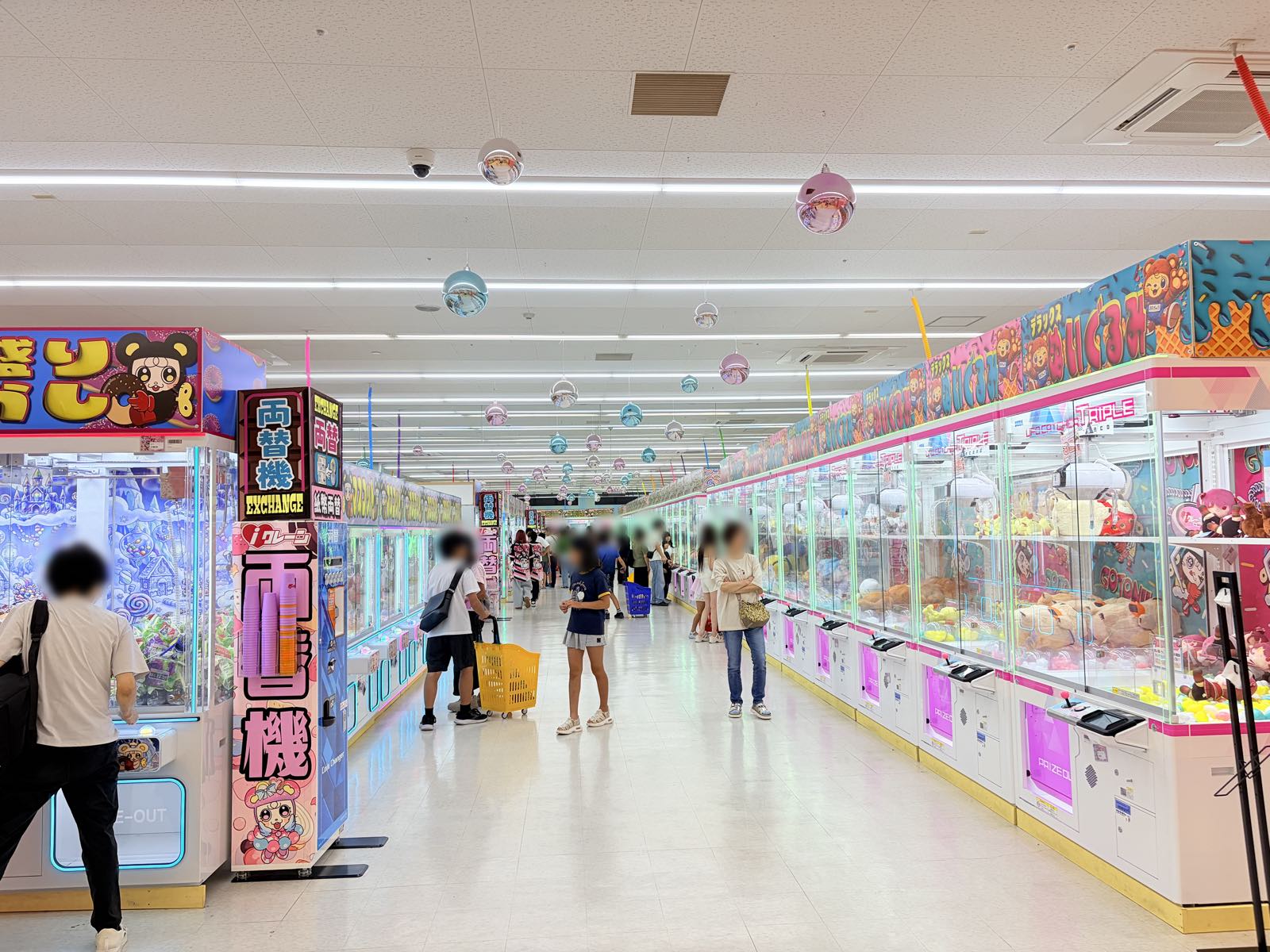 ぬいぐるみが入ったクレーンゲーム台が右側に並ぶ通路。左手前に「両替機」の縦看板が立ち、天井からパステルカラーのミラーボールが下がっている