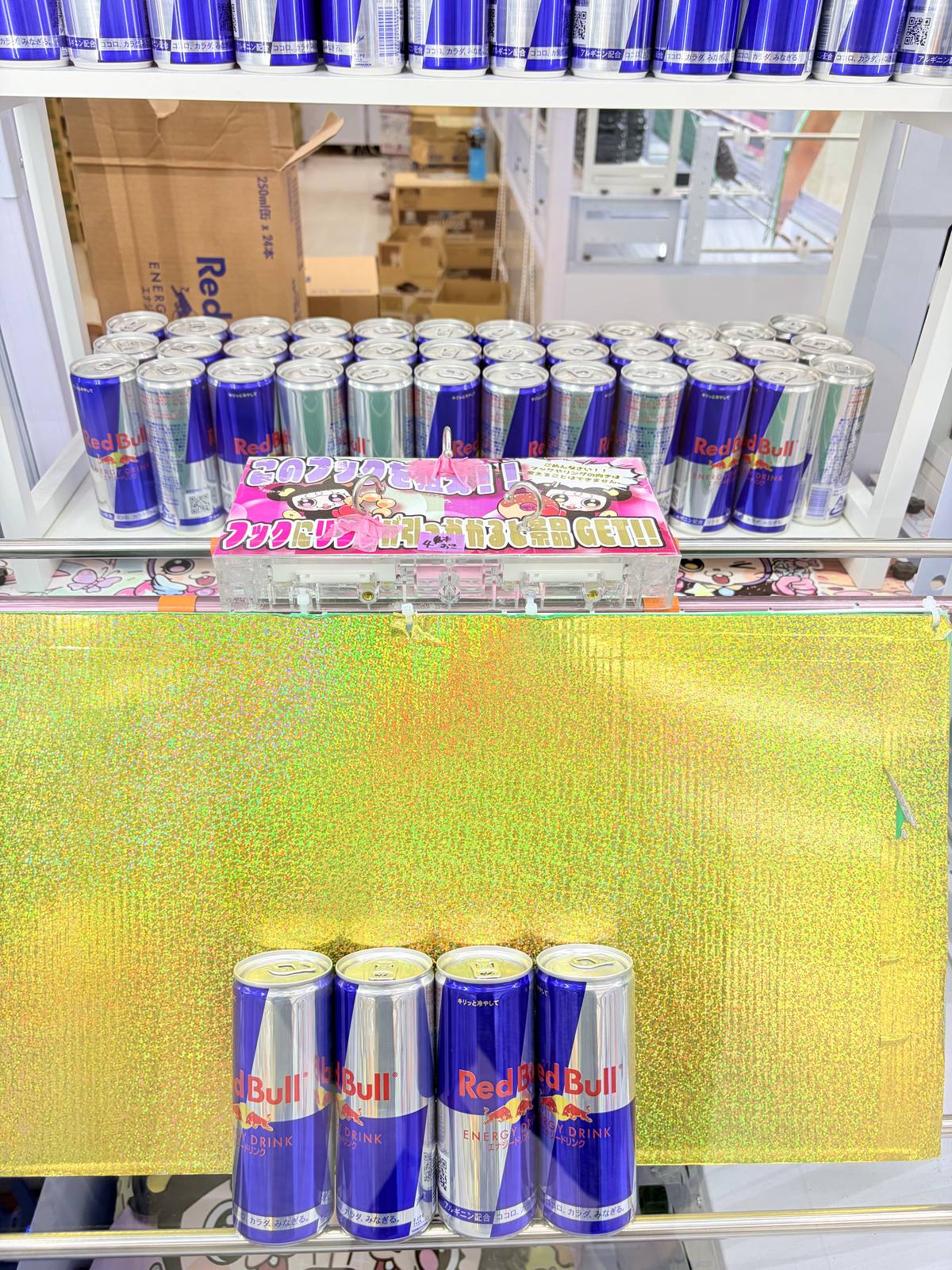レッドブルの缶が並ぶフック設定台。中央にリングが3つ下がったアクリル製の景品台があり、フックにリングを引っかけて景品を狙うことを案内するPOPが掲示されている