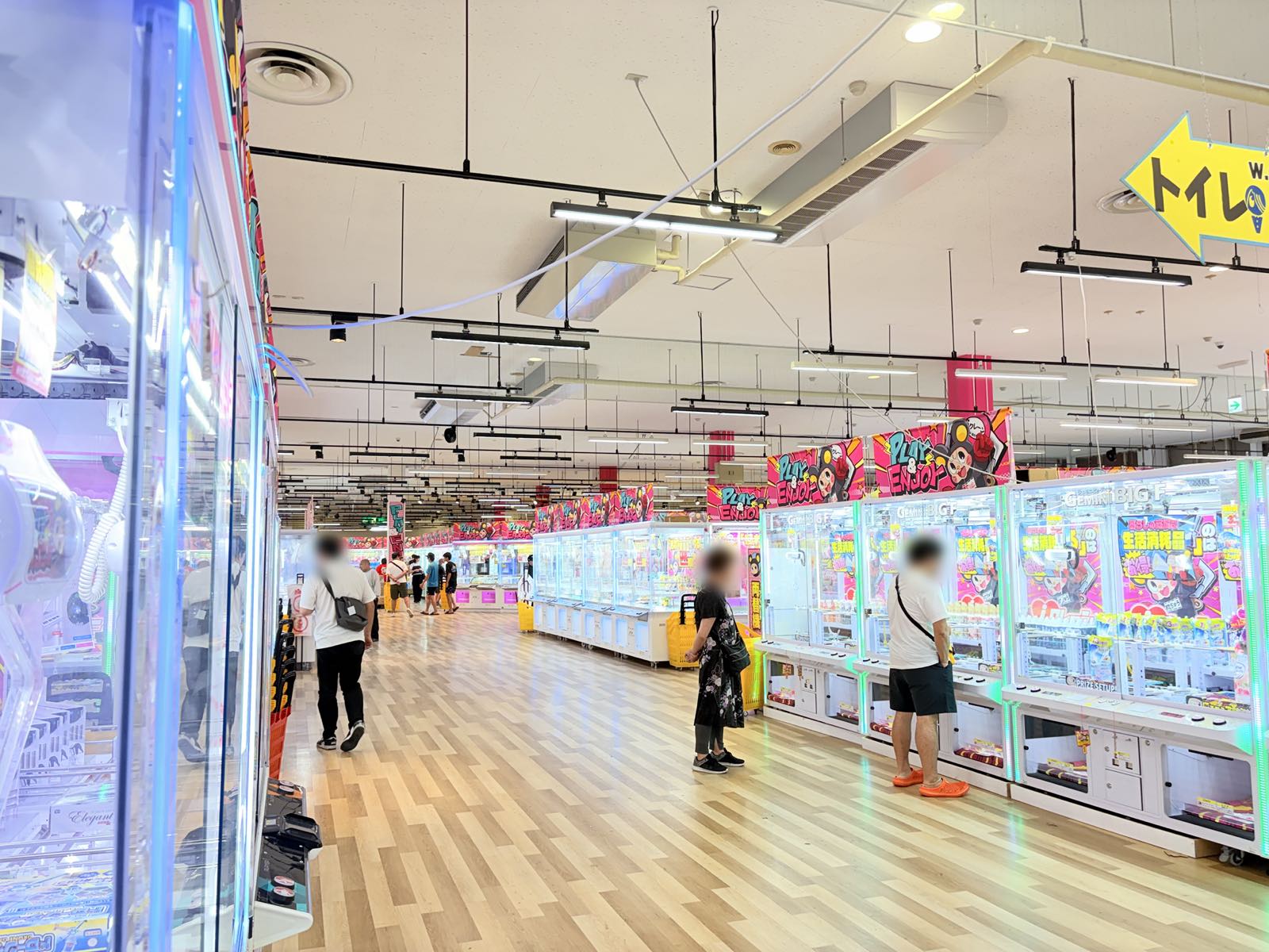 iクレーン新発田店の店内。明るい木目調の床の広い通路の両側にクレーンゲーム台がずらりと並び、天井から「PLAY & ENJOY」のバナーが下がっている