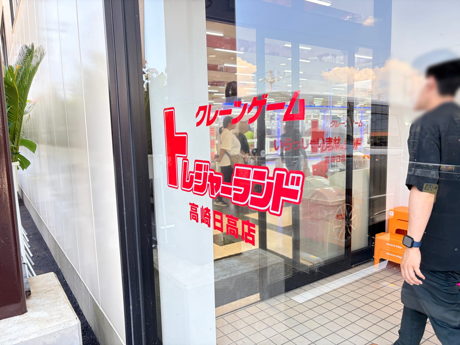 入口ガラス面に貼られた「クレーンゲーム トレジャーランド 高崎日高店」の赤いロゴ。足元には子供用のオレンジ色の踏み台が置かれている