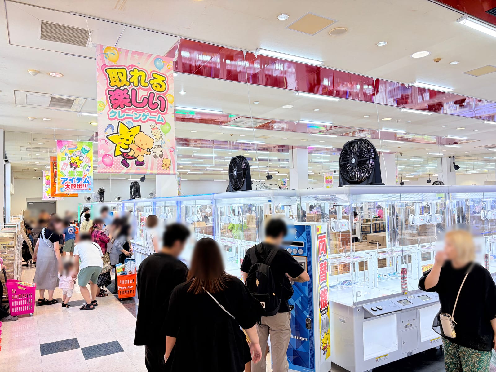 店内入口付近に吊るされた「取れる 楽しい クレーンゲーム」の大型バナーと「生活アイテム 大放出!!」ののぼり。手前に両替機、奥にクレーンゲーム台が並び、天井近くに大型送風機が設置されている