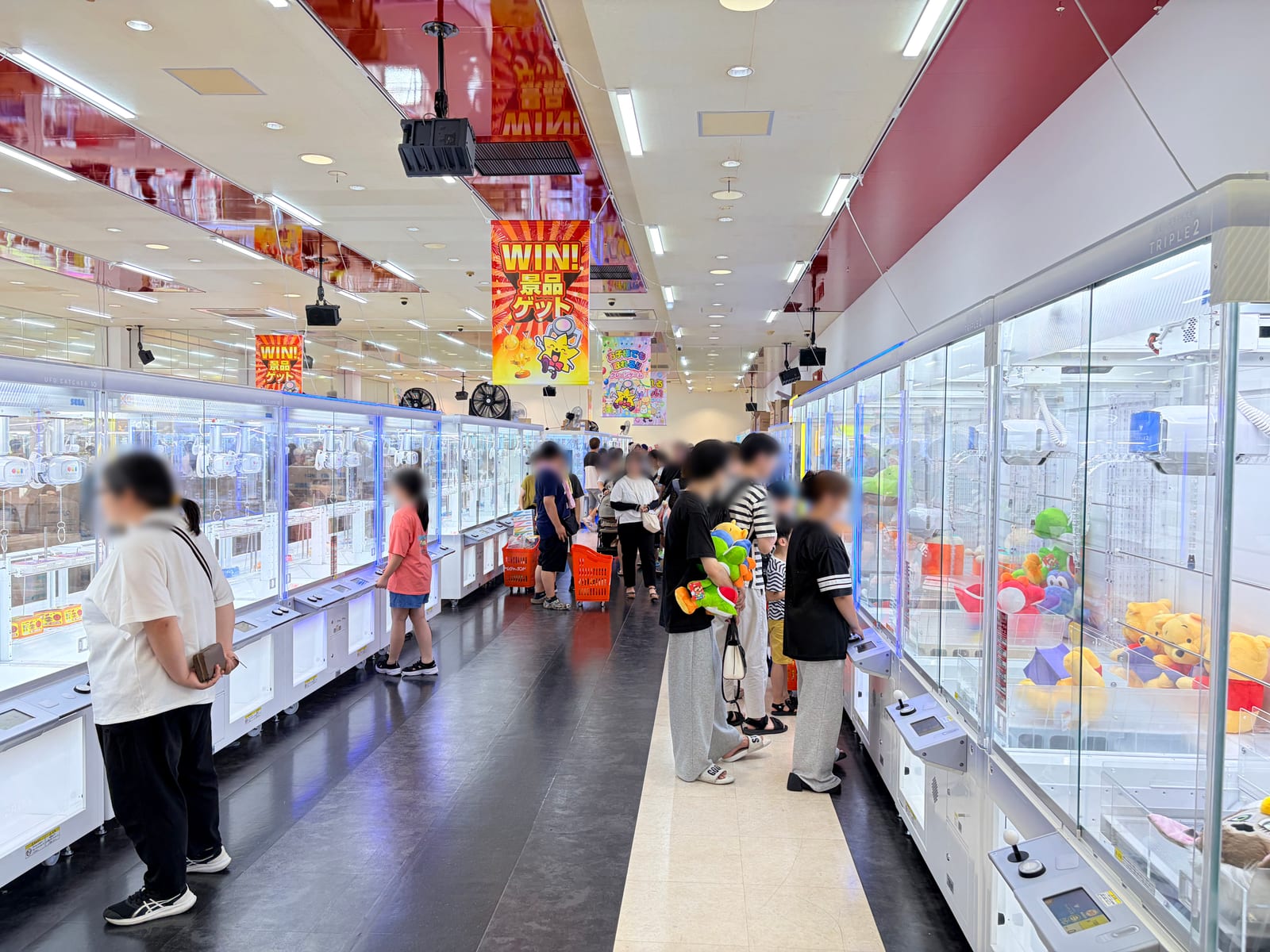 クレーンゲーム台が左右に並ぶ店内通路。天井から「WIN! 景品ゲット」のバナーが下がり、右側の台にはぬいぐるみが入っている